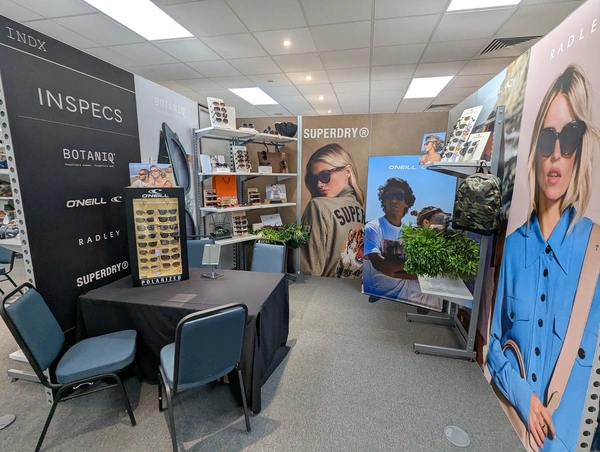 a modular stand with three 3m wide walls with colourful prints showcasing an opticians brands. a table with display of glasses and several chairs sit in front of the walls