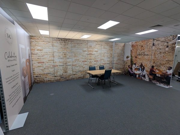 A u-shaped modular exhibition stand with images of brick walls, with a table and chair sitting infront
